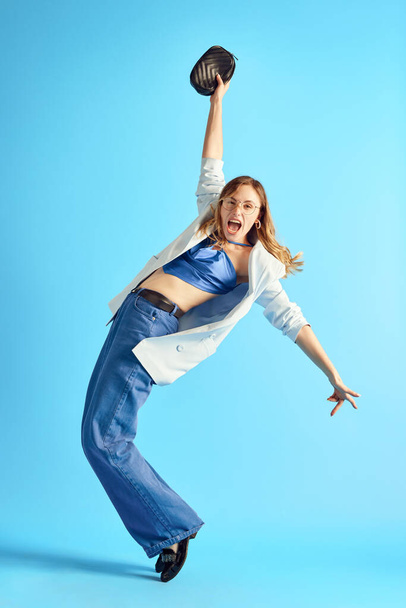 Full-length image of young woman in stylish smart casual clothes dancing, having fun against blue studio background. Concept of human emotions, youth, fashion, lifestyle, ad - Foto, Imagem