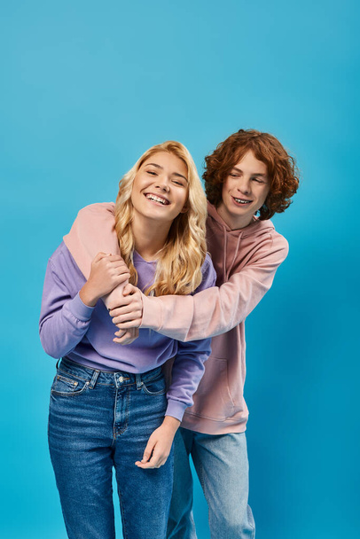 redhead teen guy embracing laughing girlfriend on blue, friendship of stylish teenagers - Photo, Image