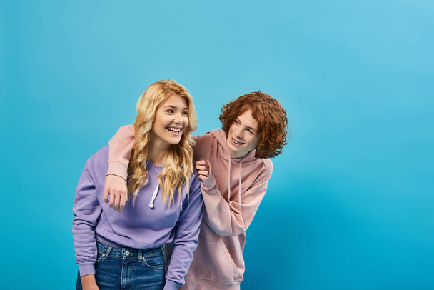 blonde teen girl laughing near redhead boyfriend, happy friends in stylish hoodies and jeans on blue - Photo, Image