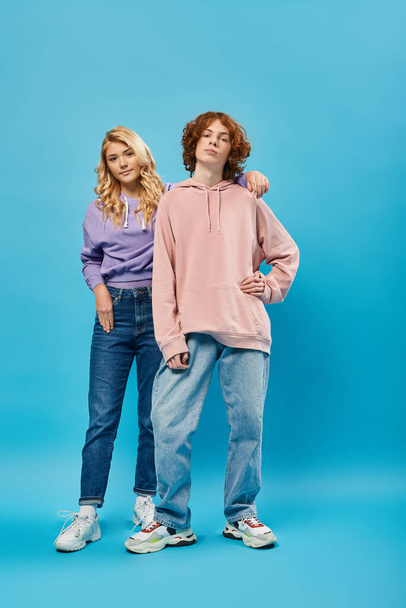 confident redhead teen guy with hand on hip near smiling blonde girlfriend on blue, stylish friends - Photo, Image