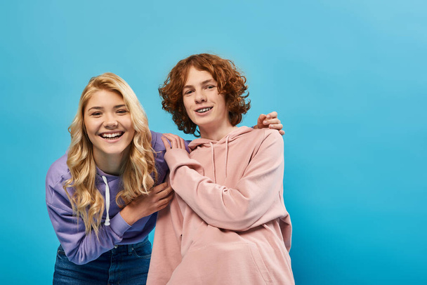 blondynka nastolatek dziewczyna przytula ruda chłopak i śmieje się z kamery na niebieski, szczęście i jedność - Zdjęcie, obraz