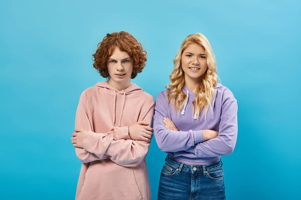 irritated teenage friends in hoodies standing with crossed arms and looking at camera on blue - Foto, afbeelding