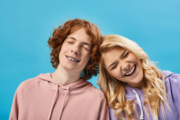 portrait of excited teenage boy and girl in hoodies laughing with closed eyes on blue, happiness - Fotoğraf, Görsel