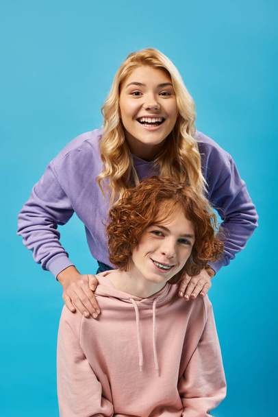 cheerful and energized blonde teen girl looking at camera near smiling redhead boyfriend on blue - Fotoğraf, Görsel