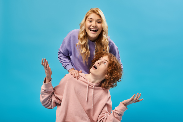 excited blonde teen girl having fun and laughing at camera near surprised redhead boyfriend on blue - Фото, изображение
