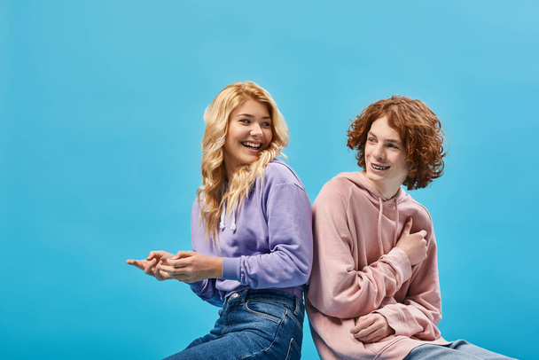 happy teenage friends in trendy outfits sitting back to back and smiling at each other on blue - Photo, Image