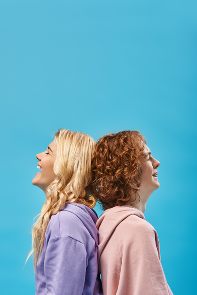 side view of excited blonde girl with redhead friend laughing with closed eyes back to back on blue - Fotoğraf, Görsel