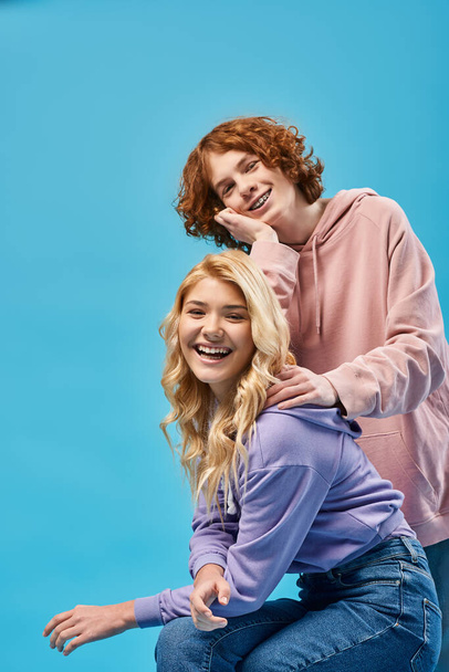 heureux rousse adolescent garçon penché sur blonde copine regarder caméra et rire sur bleu - Photo, image