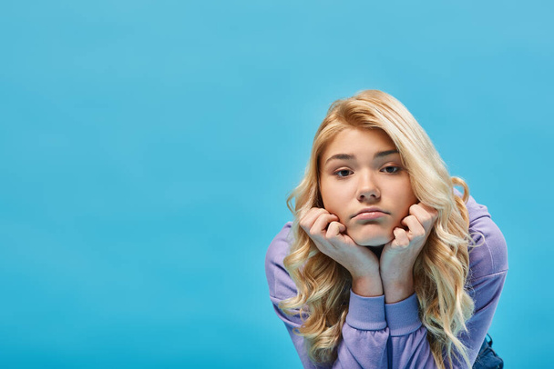 Frustriertes blondes Teenager-Mädchen im Kapuzenpulli hält die Hände vor dem Gesicht und blickt in die Kamera auf blau - Foto, Bild