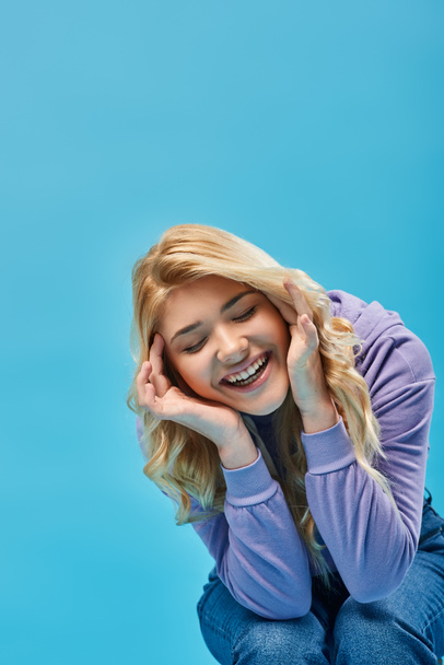 cheerful blonde teen girl in hoodie laughing with hands near face and closed eyes on blue, happiness - Photo, Image