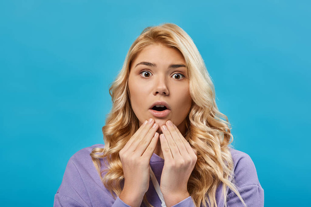astonished blonde teen girl with open mouth holding hands near chin and looking at camera on blue - Photo, Image