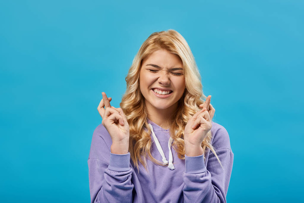 tense blonde teenage girl with closed eyes holding crossed fingers and wishing luck on blue - Photo, Image