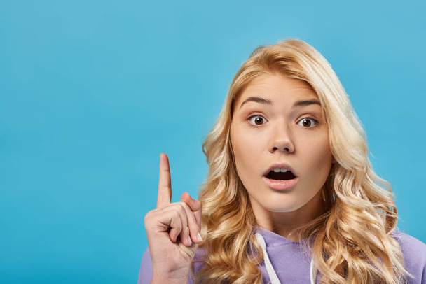 πορτρέτο της έκπληξης ξανθιά έφηβη κοπέλα με ανοιχτό στόμα δείχνει χειρονομία ιδέα στο μπλε - Φωτογραφία, εικόνα