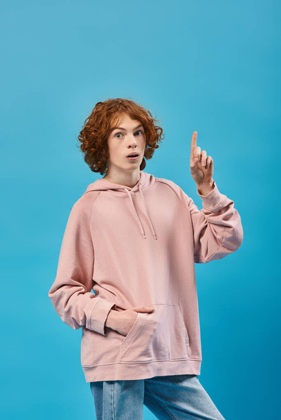 amazed redhead teen guy showing idea sign, holding hand in pocket and looking at camera on blue - Photo, Image