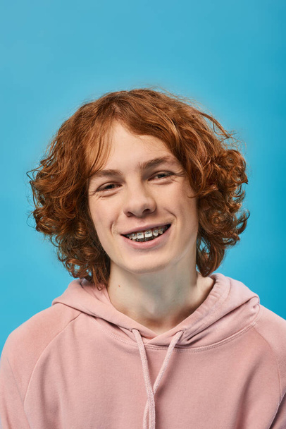 портрет веселого подростка с рыжими волнистыми волосами и брекетами, улыбающимися в камеру на голубом, счастье - Фото, изображение