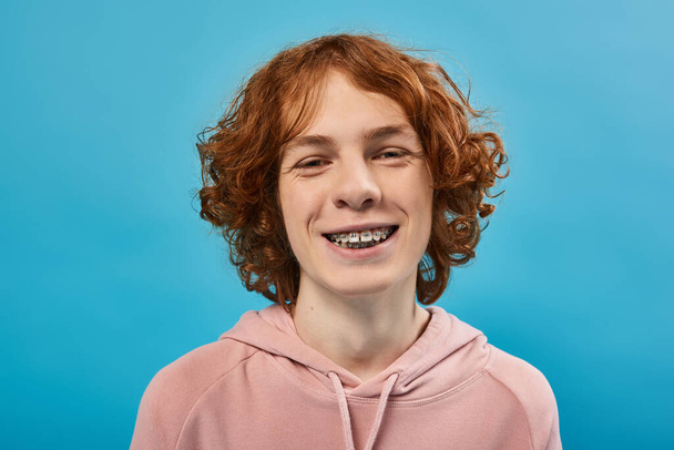 carefree and joyful teenage guy with red wavy hair and braces looking at camera on blue, portrait - Photo, Image