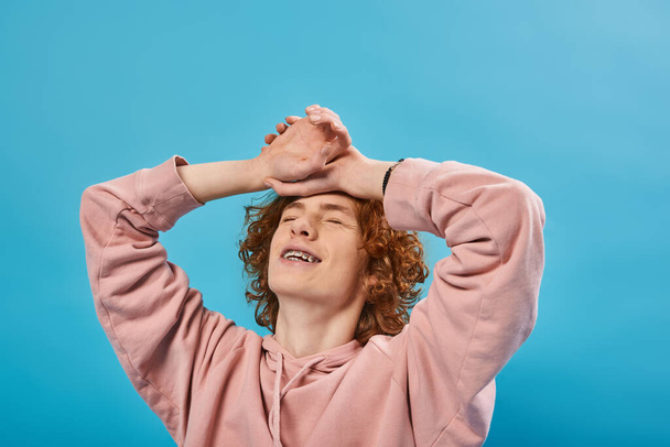 positive, dreamy redhead teen guy in hoodie standing with closed eyes and hands on forehead on blue - Foto, Bild