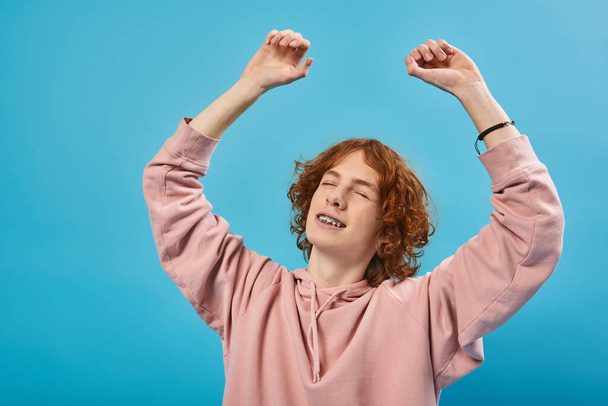 smiling and dreamy redhead teen guy in hoodie standing with closed eyes and raised hands on blue - Photo, Image