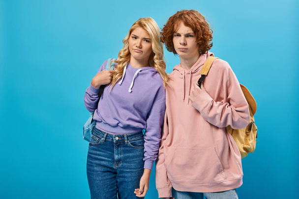 displeased teenage classmates standing with school backpacks and looking at camera on blue - Zdjęcie, obraz