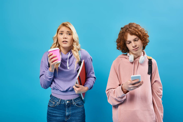 zaniepokojona blondynka nastolatka dziewczyna z papierowym kubkiem w pobliżu uśmiechnięty przyjaciel ze smartfonem i słuchawki na niebiesko - Zdjęcie, obraz