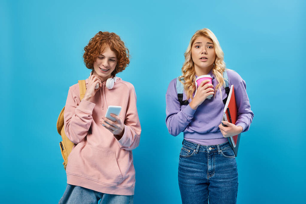 nervosa ragazza adolescente con tazza di carta guardando la fotocamera vicino amico positivo utilizzando smartphone su blu - Foto, immagini