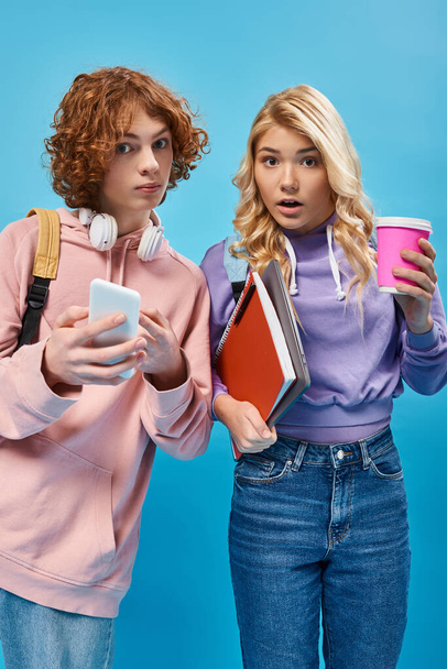 изумленные подростки с рюкзаками, наушниками и цифровыми устройствами, смотрящие в камеру на голубом - Фото, изображение