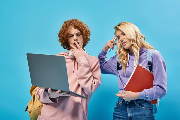 geschokt en attent tiener klasgenoten met rugzakken kijken naar laptop op blauw - Foto, afbeelding