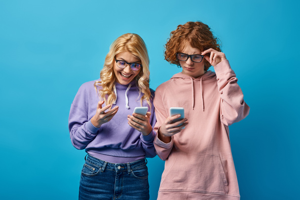 χαρούμενη ξανθιά έφηβη κοπέλα με γυαλιά κοιτάζοντας το smartphone κοντά στοχαστική κοκκινομάλλα φίλη στο μπλε - Φωτογραφία, εικόνα