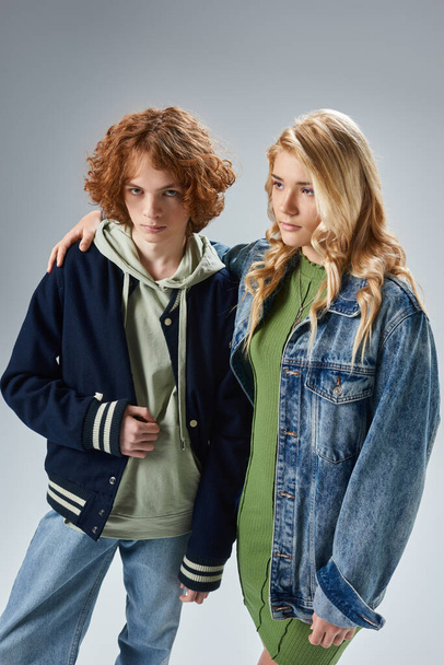confident, trendy redhead teen boy looking at camera near blonde girlfriend on grey, youth fashion - Photo, Image