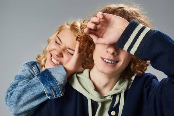 cheerful blonde teenage girl with closed eyes obscuring face of stylish redhead friend on grey - Fotografie, Obrázek
