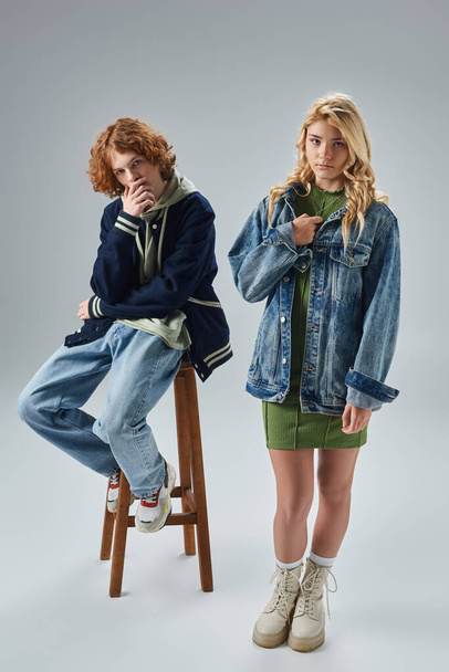 zrzky a stylový teen chlap sedí na vysoké stoličce v blízkosti blondýny přítelkyně na šedé, dospívající móda - Fotografie, Obrázek