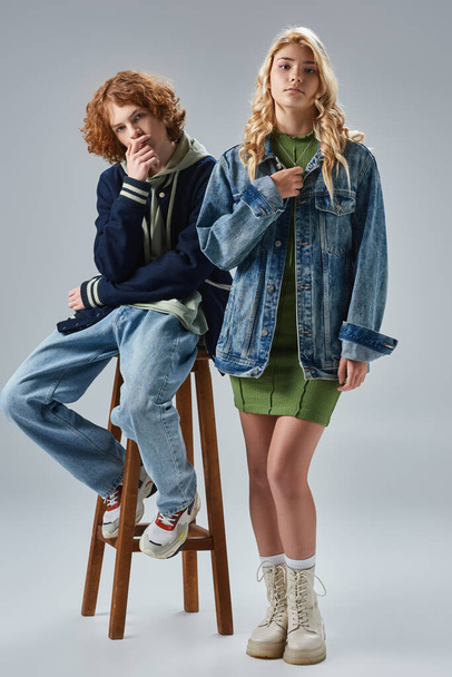 blonde teen girl in trendy outfit looking at camera near redhead boy on high stool on grey - Foto, imagen