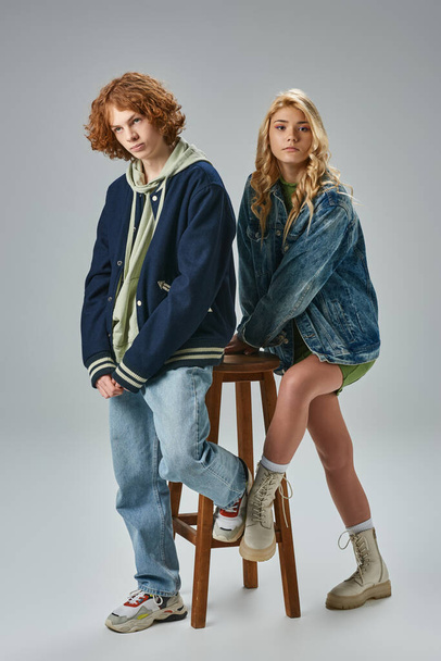 full length of teenage boy and girl in trendy casual attire posing near high stool on grey - 写真・画像