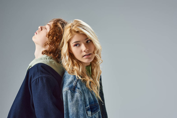 redhead and blonde teen models standing back to back while posing in trendy casual attire on grey - Photo, Image