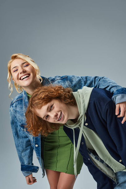 laughing blonde teen girl embracing stylish redhead boyfriend while having fun in studio on grey - Photo, Image