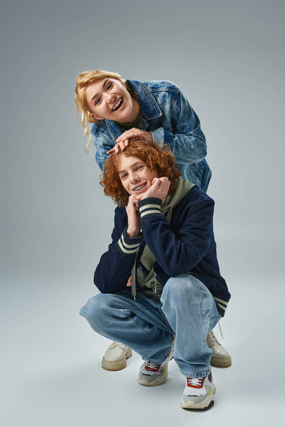 excited youthful girl laughing near redhead stylish guy sitting on haunches on grey, teen fashion - Fotografie, Obrázek