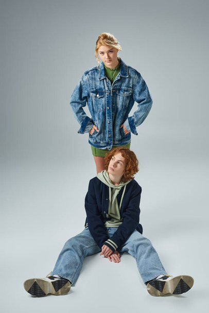 confident teen girl with hands in pockets looking at camera near trendy redhead guy sitting on grey - Φωτογραφία, εικόνα