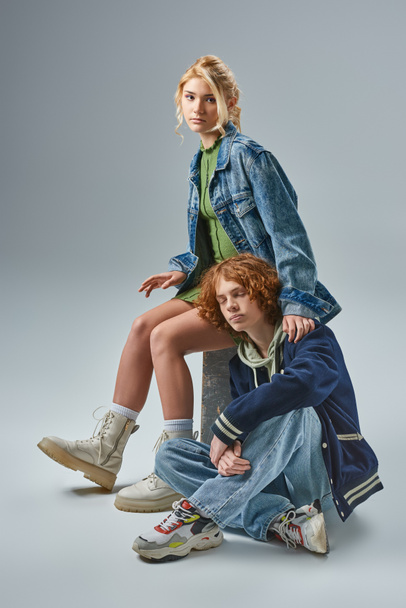 blonde teen girl in trendy wear looking at camera near redhead guy sitting with closed eyes on grey - Фото, зображення