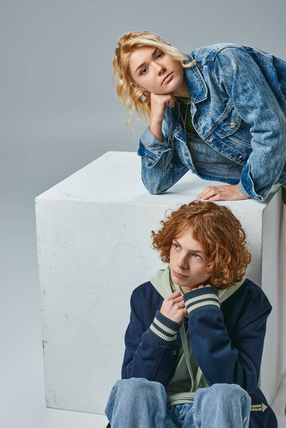 blondes Teenie-Mädchen in stilvoller Kleidung schaut in die Kamera in der Nähe von Rotschopf Kerl sitzt in der Nähe Würfel auf grau - Foto, Bild