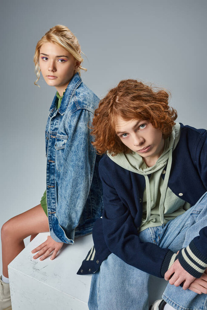 couple of teen models in trendy casual attire sitting on white cube and looking at camera on grey - Zdjęcie, obraz