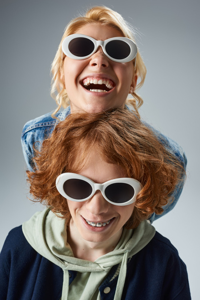 excited blonde teen girl in trendy sunglasses laughing near redhead boyfriend on grey, youth fashion - Foto, Bild
