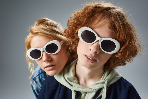 πορτρέτο του κοκκινομάλλα έφηβος τύπος και η φίλη σε μοντέρνα γυαλιά ηλίου κοιτάζοντας κάμερα σε γκρι - Φωτογραφία, εικόνα