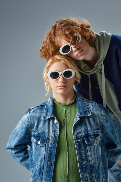 coppia di modelli adolescenti in abbigliamento casual alla moda e occhiali da sole in posa e guardando la fotocamera sul grigio - Foto, immagini