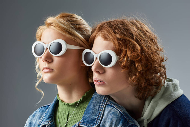 redhead teenage boy and blonde girlfriend in sunglasses and stylish casual clothes posing on grey - Photo, Image