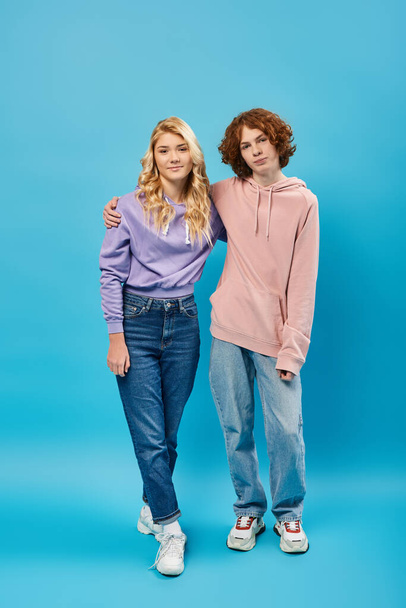 šťastný dospívající přátelé v mikinách a džínách při pohledu na kameru, zatímco stojí na modré, plné délce - Fotografie, Obrázek