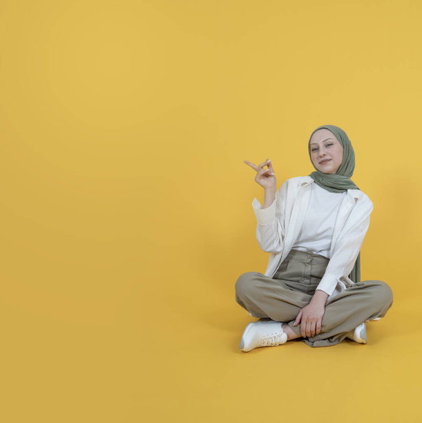コピースペースを脇に置いて,ヒジャブの座席の床に完全なボディ白人イスラム教徒の女性がコピースペースを指している. プロダクト表示を推薦する表示. 黄色い隔離されたスタジオの背景にカメラを見る.  - 写真・画像