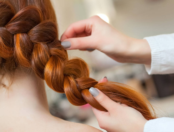 Beautiful girl with long red hair, hairdresser weaves a braid, in a beauty salon. Professional hair care and creating hairstyles. - Photo, Image