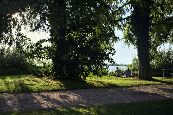 Paar sitzt am Seeufer unter Bäumen in einem Park bei Sonnenuntergang - Foto, Bild