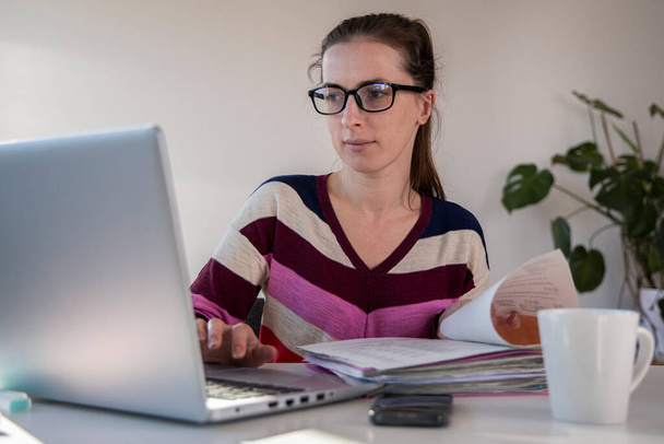 Mujer joven en gafas mirando los papeles mientras está sentada en un ordenador portátil. - Foto, Imagen