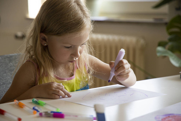 klein kind blond meisje tekent terwijl zitten aan de tafel. - Foto, afbeelding